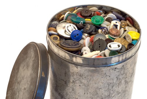 The image shows a round metal container, possibly a trash or storage bin, filled with a diverse collection of plastic buttons of various sizes, shapes, and colors, including blue, white, black, brown, yellow, and green. The container's exterior appears worn with visible scratches and a slightly tarnished surface, indicating previous use. Its lid, made of metal matching the container, is partially visible leaned against the side, suggesting the bin has been recently opened or is in the process of being filled or emptied. The buttons inside appear to be made of plastic with some having textured or shiny surfaces, and they are loosely piled, with some overlapping each other. The environment context is neutral with a clean background, emphasizing the focus on the container and its contents. The overall scene relates to rubbish or waste collection, aligning with the services provided by Business Waste Removal Camden Town, which handles the disposal and removal of household or commercial waste materials generally found in UK towns and cities.
