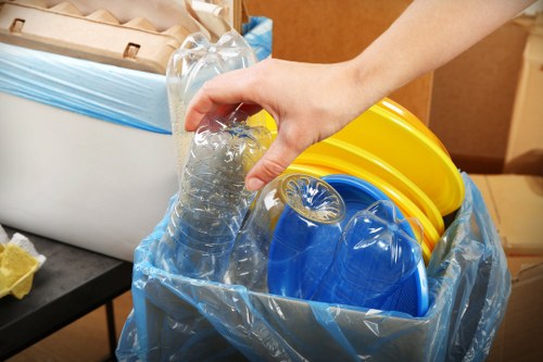 A close-up view of a person's hand holding a clear plastic bottle above a large blue recycling bin lined with a transparent plastic bag. Inside the bin are several brightly colored plastic items, including yellow and blue buckets and containers. The background shows part of a domestic or commercial space with a wooden surface and some packaging or cardboard boxes nearby, suggesting an indoor area designated for waste sorting or collection. The image emphasizes the process of sorting recyclable plastic waste, aligning with the services offered by Business Waste Removal Camden Town for efficient rubbish collection and sustainable waste management in the local area.