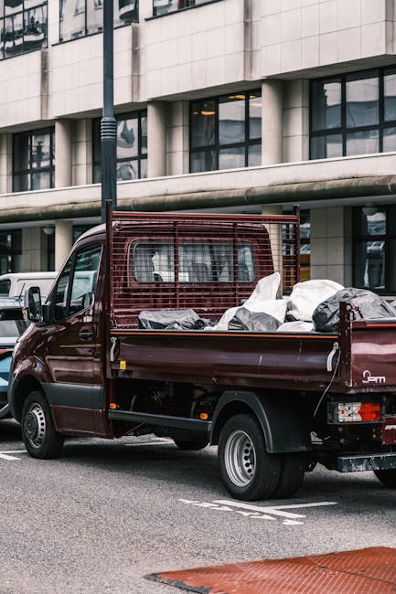 A small burgundy flatbed truck parked on a city street in front of a modern office building with large windows. The truck's cargo area is filled with assorted rubbish, including black and white bags, 
